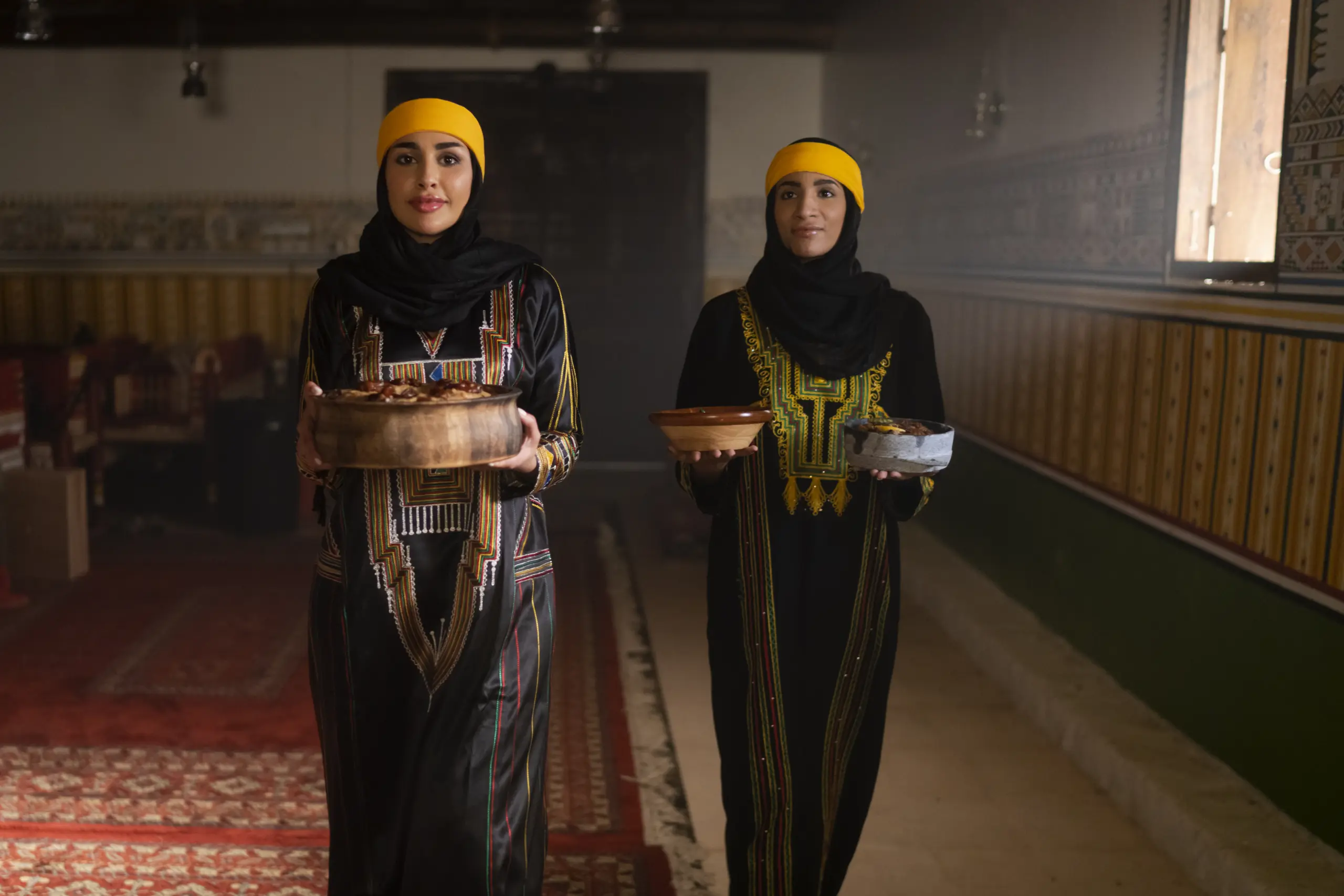 Two women wearing traditional Qahtani clothing carrying dishes o